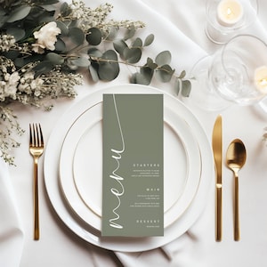 Puede incluir: Una tarjeta de menú verde con fuente blanca de escritura que dice "menú" y enumera "Entrantes", "Plato principal" y "Postre". La tarjeta está sobre un mantel blanco con un plato blanco, cubiertos de oro y una vela encendida.