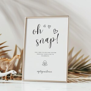 Peut inclure: Un panneau encadré avec les mots "oh snap!" en écriture noire, décoré de coeurs. Le panneau comprend également du texte sur un géo-filtre Snapchat et un hashtag. Deux dinosaures jouets se tiennent devant le panneau.