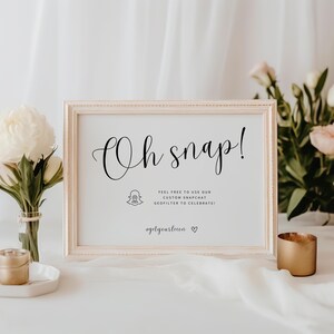 Peut inclure: Un panneau encadré avec les mots "Oh snap!" en écriture noire élégante. Le panneau comprend le logo Snapchat et du texte encourageant l'utilisation d'un géo-filtre personnalisé. La scène est décorée de fleurs et d'un bougeoir doré.