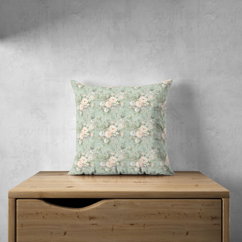 K&ouml;nnte beinhalten: Ein hellgr&uuml;nes Kissen mit einem Blumenmuster mit wei&szlig;en und rosa Rosen. Das Kissen liegt auf einer Holzoberfl&auml;che.