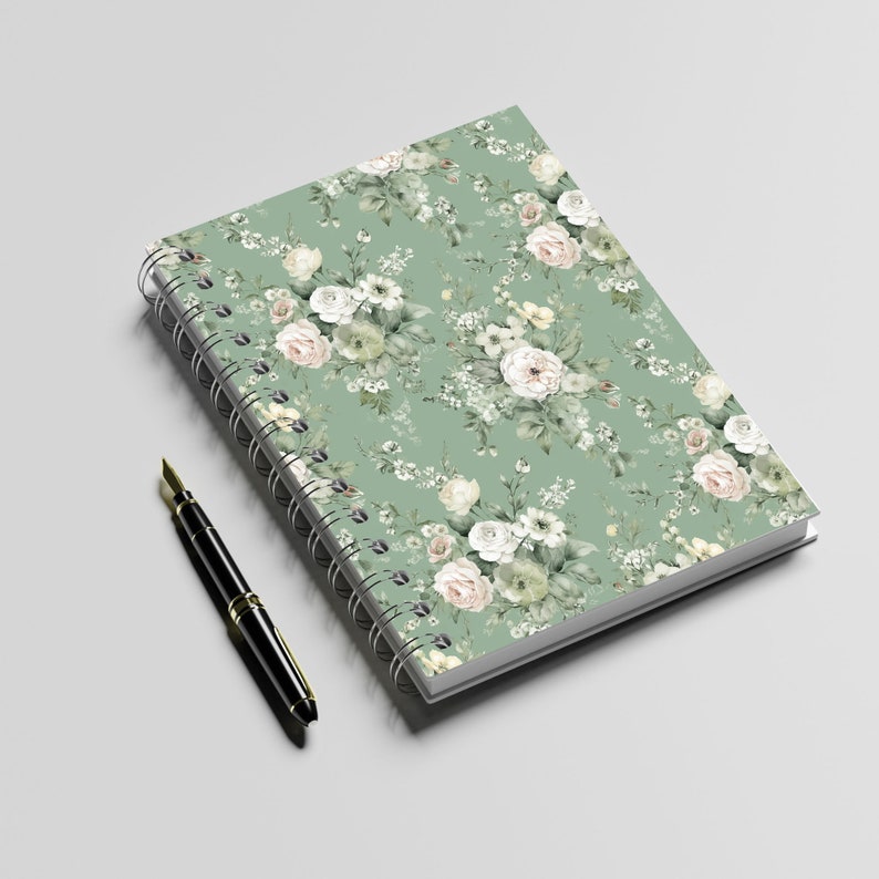 K&ouml;nnte beinhalten: Ein gr&uuml;nes Spiralnotizbuch mit einem wei&szlig;en Blumenmuster. Das Notizbuch liegt flach auf einer wei&szlig;en Oberfl&auml;che mit einem schwarzen Stift daneben.