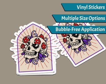 Puede incluir: Una pegatina de vinilo con una calavera con setas rojas y blancas que le salen de la cabeza. La calavera está rodeada de hojas moradas y un patrón de rayos de sol rojos y blancos. La pegatina está disponible en varios tamaños y tiene una aplicación sin burbujas.