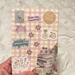 May include: A decorative card with a pink and yellow gingham pattern. The card features stickers with floral and book themes, including the phrases "hello sunshine", "shhh! i'm reading", and "little miss homebody".