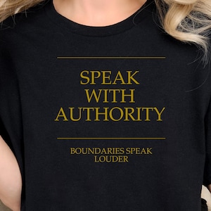 May include: Black t-shirt with gold text that reads "SPEAK WITH AUTHORITY" above "BOUNDARIES SPEAK LOUDER". The shirt is a crew neck style and is made of a soft material.