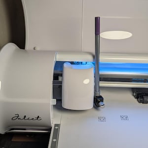 以下が含まれることがあります： 紫と銀色の切削工具を備えた白いJuliet切断機。機械には青色のライトと、小さな楕円形のライトが付いた白い円筒形のコンポーネントがあります。「Juliet」の文字が機械に書かれています。