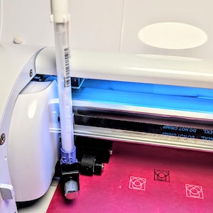 May include: A white vinyl cutting machine with a blue light illuminating the cutting area. A white pen-like tool is positioned near the blade. The machine is cutting a pink sheet of material with a repeating design.