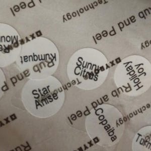 May include: A close-up of several small, circular stickers with various text labels. The stickers are white with black text, including phrases like "Sunny Citrus," "Star Anise," and "Holiday Joy." The background is a translucent material with additional text.