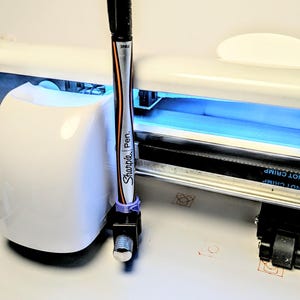 以下が含まれることがあります： 黒いSharpieペンが、機械に取り付けられた紫色のホルダーに保持されています。ペンは、オレンジと黒のアクセントが付いたシルバーのバレルを持っています。機械は白で、青色のライトが付いています。
