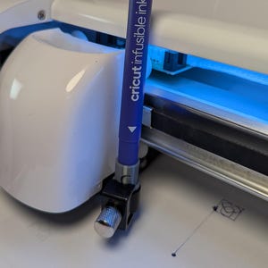 以下が含まれることがあります： 青いCricut Infusible Inkペンを使用しているCricutマシンのクローズアップ。ペンは黒とシルバーのクランプで固定されています。機械は白で、青色のライトが付いています。