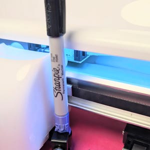May include: A black and white Sharpie permanent marker is held in a purple and black holder. The marker is positioned in front of a white machine with a blue light. The machine is on a pink surface.