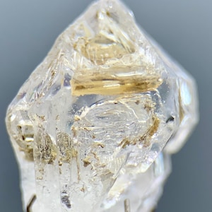 Puede incluir: Primer plano de un cristal de cuarzo transparente con inclusiones internas y una forma puntiaguda y facetada. El cristal tiene un aspecto translúcido con áreas de color marrón claro y beige. El cuarzo está montado sobre un fino soporte metálico.