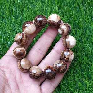 Pode incluir: Uma pulseira de contas com grandes contas redondas em tons de castanho e creme. As contas têm um acabamento polido e brilhante e um padrão listrado. A pulseira é exibida em um fundo de grama verde.