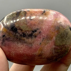 May include: A polished, oval-shaped rhodonite gemstone. The stone displays a mix of pink, black, and brown hues, with a smooth, glossy surface. The gemstone is held in a hand, showcasing its natural beauty and unique color variations.
