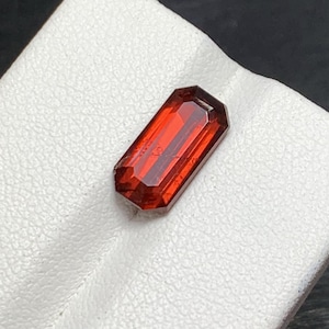 May include: A rectangular, faceted gemstone with a deep red hue. The gem has a polished surface and a symmetrical, elongated shape. The gemstone is displayed on a white, textured surface.