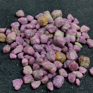 Może przedstawiać: Stos surowych kamieni szlachetnych rubinu. Kamienie różnią się rozmiarem i kształtem, wykazując gamę różowych i czerwonofioletowych odcieni. Niektóre kamienie mają lekko brązowawy odcień, a inne wykazują zielone odcienie. Kamienie szlachetne są rozrzucone na czarnej powierzchni.