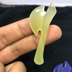 May include: A translucent, pale yellow axe-shaped pendant. The pendant has a curved handle and a rounded head with a notch. The stone has a smooth, polished surface and is held by a hand against a dark background.