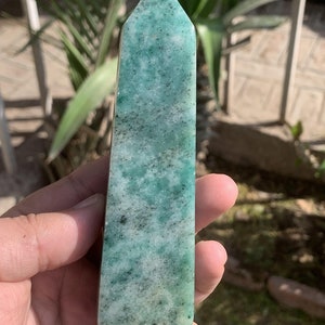 Può includere: Un obelisco di cristallo rettangolare e lucido con una parte superiore a punta. Il cristallo è di un colore verde acqua screziato con macchie scure. L'obelisco è tenuto in mano, con uno sfondo sfocato di fogliame verde.