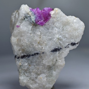 May include: A rough, uncut gemstone specimen featuring a vibrant, fuchsia-colored crystal cluster. The stone is set in a matrix of white and gray rock, with dark, linear inclusions. The overall texture is coarse and natural, showcasing the raw beauty of the mineral.