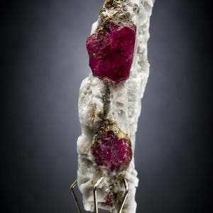 Puede incluir: Un espécimen mineral vertical que presenta dos cristales de rubí de color rojo magenta intenso incrustados en una matriz texturizada de color blanco hueso. Incluye pequeñas motas de pirita metálica dorada y se apoya en un soporte de metal.
