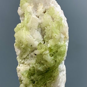 May include: A mineral specimen featuring a cluster of light green crystals on a white matrix. The crystals have a prismatic shape and are clustered together. The background is a solid gray color.