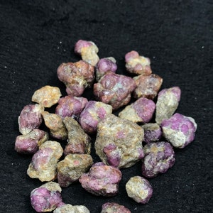 Könnte beinhalten: Eine Sammlung roher, ungeschliffener Rubin-Edelsteine. Die Steine zeigen eine Mischung aus tiefroten und violettlichen Farbtönen mit einer rauen, strukturierten Oberfläche. Die Edelsteine sind auf einem dunklen Hintergrund verstreut.