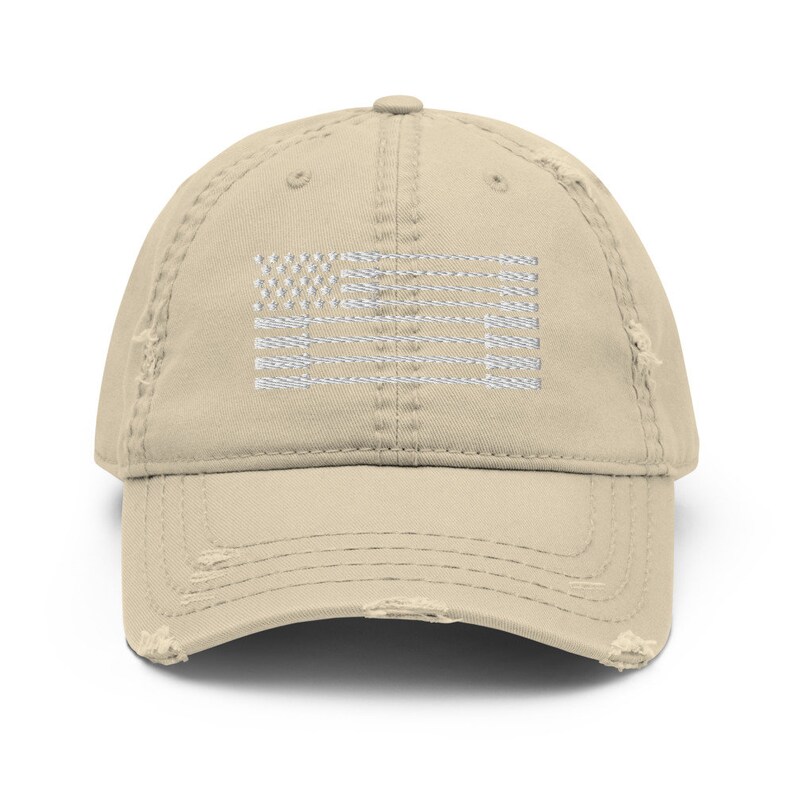 May include: A distressed tan baseball cap featuring a white embroidered American flag design. The cap has a curved brim and a worn, vintage look with distressed edges. This hat is a stylish accessory for casual wear.