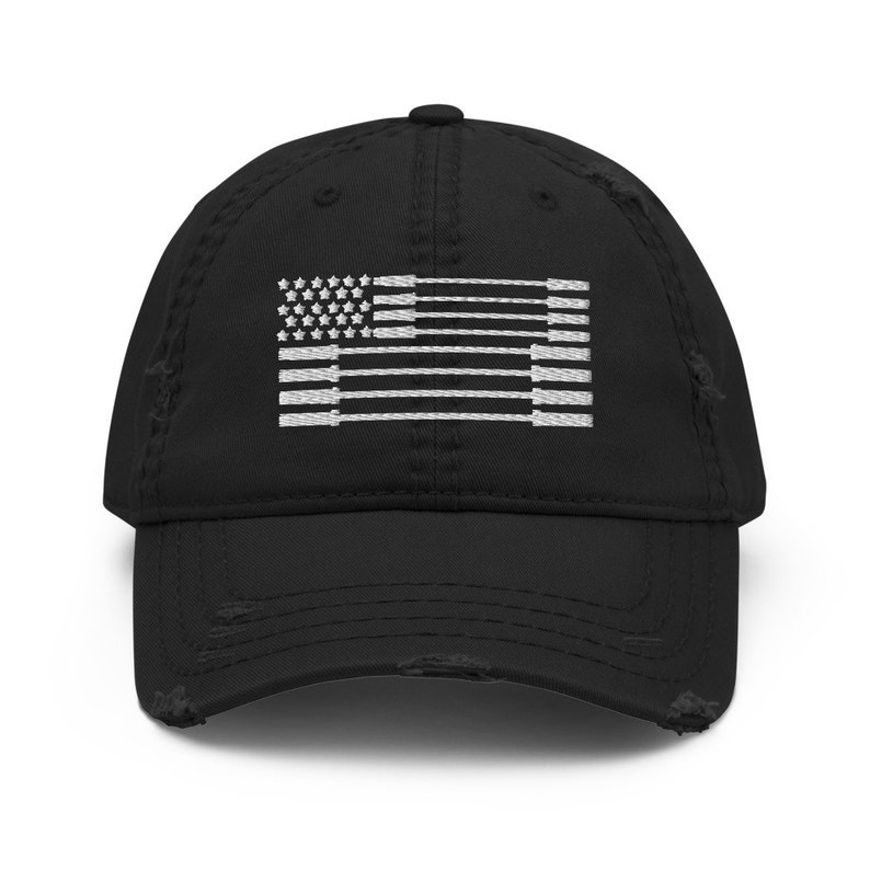 May include: Black distressed baseball cap with a white embroidered flag design. The flag features stars and stripes, with the stripes appearing as weightlifting barbells. The cap has a curved brim and a worn, vintage look.