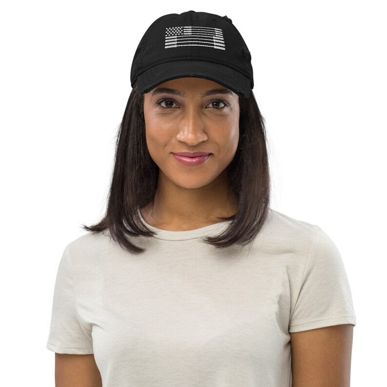 May include: Black distressed baseball cap with a white graphic of the American flag. The cap is worn by a person with dark hair and a light-colored t-shirt. The cap has a curved brim and an adjustable strap.