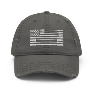 May include: A distressed gray baseball cap featuring a white embroidered design of the American flag. The flag design incorporates weightlifting barbells in place of the stripes. The cap has a pre-worn look with distressed edges.