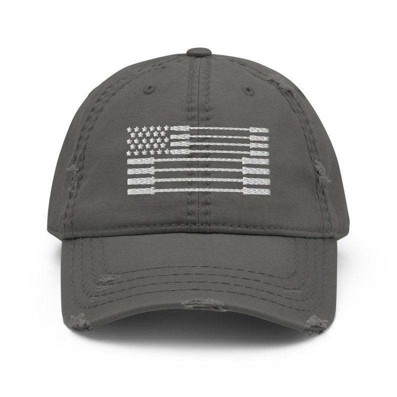 May include: A distressed gray baseball cap featuring a white embroidered design of the American flag. The flag design incorporates weightlifting barbells in place of the stripes. The cap has a pre-worn look with distressed edges.