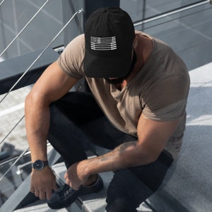 May include: Black baseball cap with a white graphic of a flag design. The cap is made of a soft material and has a curved brim. The cap is worn by a person sitting on a set of stairs.