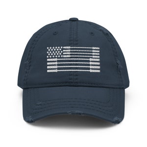 May include: A distressed navy blue baseball cap featuring a white embroidered flag design. The flag incorporates stars and stripes, with a worn, vintage aesthetic. The cap has a curved brim and adjustable strap.
