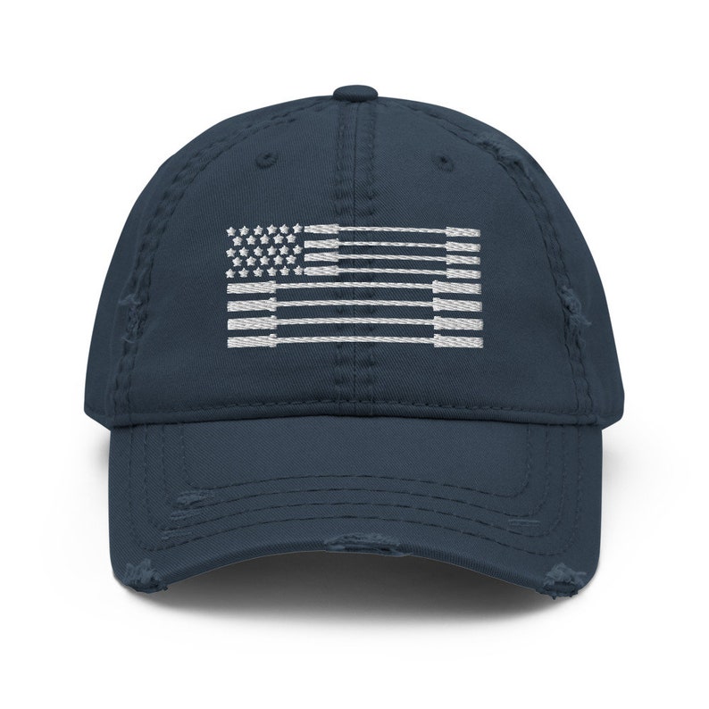 May include: A distressed navy blue baseball cap featuring a white embroidered flag design. The flag incorporates stars and stripes, with a worn, vintage aesthetic. The cap has a curved brim and adjustable strap.