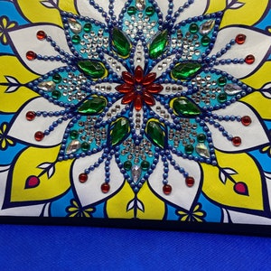 May include: A colorful mandala design with blue, green, red, and white rhinestones. The design is on a white background with a yellow and blue border.