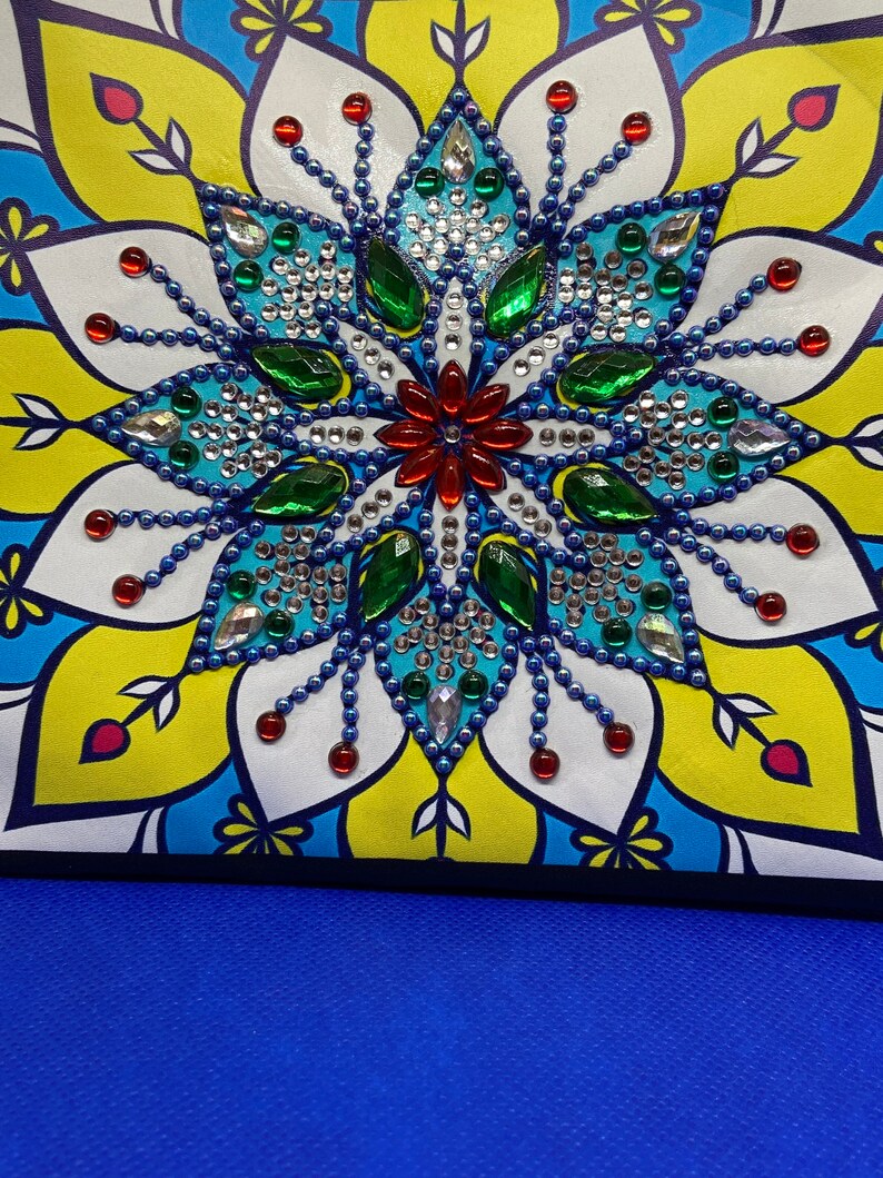 May include: A colorful mandala design with blue, green, red, and white rhinestones. The design is on a white background with a yellow and blue border.