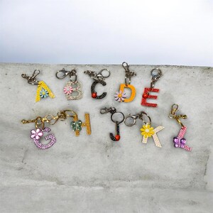 May include: A collection of letter-shaped keychains. Each letter is adorned with colorful rhinestones and a small flower accent. The keychains are in various colors, including yellow, pink, red, and green, and are attached to silver-tone clasps.