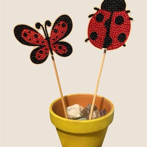 May include: A yellow painted terracotta pot holds decorative butterfly and ladybug garden stakes. The butterfly and ladybug are red and black, with a sparkling rhinestone design. The pot contains small rocks.