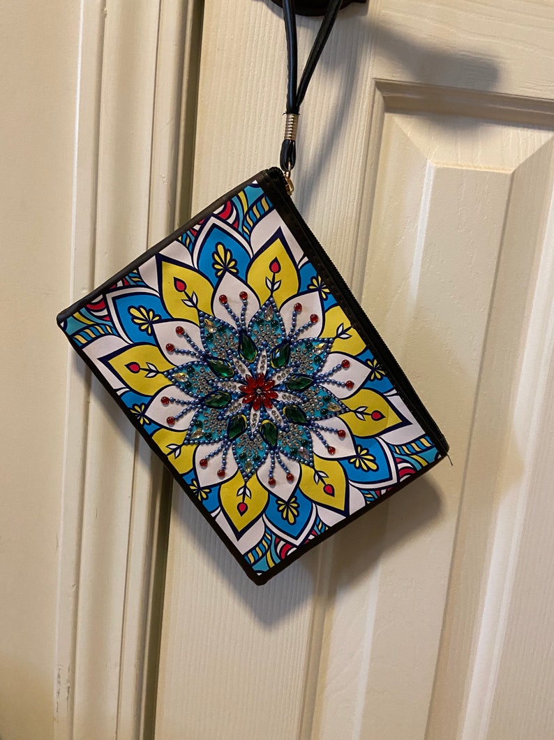 May include: A white zippered pouch with a colorful mandala design featuring blue, yellow, red, and green colors. The mandala has a beaded center with a black border around the pouch.