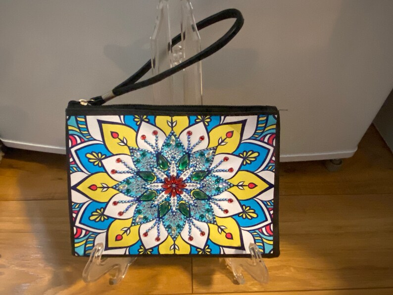 May include: A black zippered clutch with a colorful mandala design featuring a large blue, green, and red rhinestone flower in the center. The clutch has a black strap.