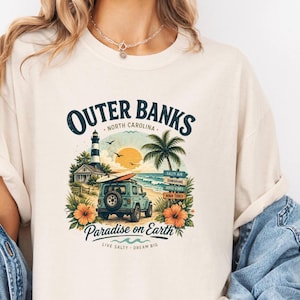 May include: Cream-colored t-shirt featuring a colorful Outer Banks design. The graphic includes a lighthouse, palm tree, beach scene, and a jeep with a surfboard. Text reads "Outer Banks, North Carolina" and "Paradise on Earth."