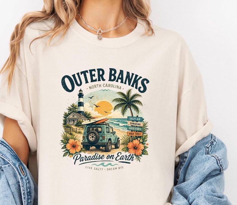 May include: Cream-colored t-shirt featuring a colorful Outer Banks design. The graphic includes a lighthouse, palm tree, beach scene, and a jeep with a surfboard. Text reads "Outer Banks, North Carolina" and "Paradise on Earth."