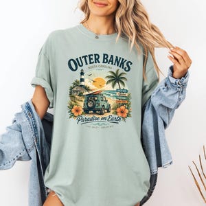 May include: A light green t-shirt with a graphic featuring the text "Outer Banks North Carolina" and a beach scene with a lighthouse, palm tree, and a jeep. The shirt also has the text "Paradise on Earth".