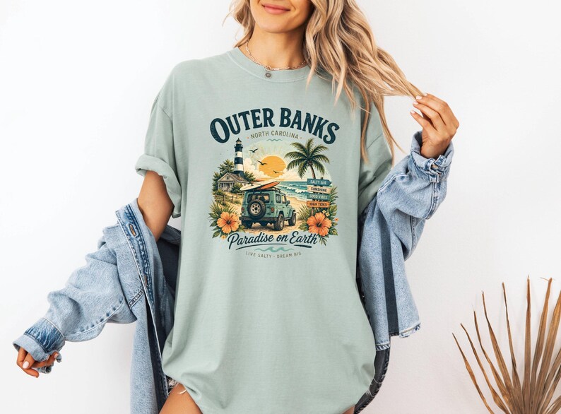 May include: A light green t-shirt with a graphic featuring the text "Outer Banks North Carolina" and a beach scene with a lighthouse, palm tree, and a jeep. The shirt also has the text "Paradise on Earth".