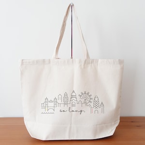 May include: A white canvas tote bag featuring a black line drawing of the London skyline, including Big Ben, the London Eye, and Tower Bridge. The text "so long." is printed below the skyline.