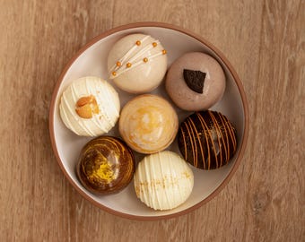 May include: A round bowl containing seven spherical chocolate treats with various decorations, including white chocolate drizzles, gold sprinkles, a dark cookie piece, and marbled brown and yellow patterns, arranged in a circular formation.
