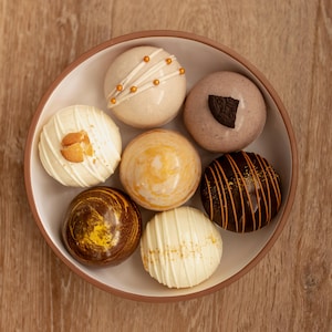 May include: A round bowl containing seven spherical chocolate treats with various decorations, including white chocolate drizzles, gold sprinkles, a dark cookie piece, and marbled brown and yellow patterns, arranged in a circular formation.