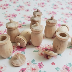 May include: A collection of small, unfinished wooden jars with matching lids. Each jar features a rounded body and a circular top with a small knob handle. The natural light wood grain is visible on the smooth surfaces of these miniature containers.