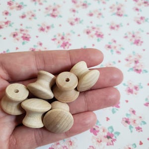 May include: A collection of small, unfinished light-colored wooden spools with a concave center and a central hole. These wooden items feature a smooth, natural grain texture and a flared top and bottom design, presented in a cluster.