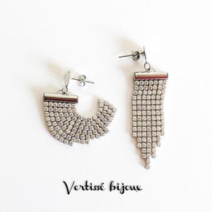 Può includere: Un paio di orecchini color argento con un design scintillante di strass. Un orecchino è a forma di semicerchio, l'altro è lungo e pendente. Il testo "Vertissé bijoux" è in corsivo.