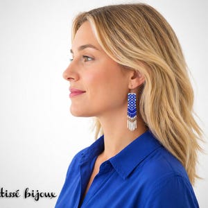 Puede incluir: Pendientes largos colgantes con un patrón geométrico en cuentas azules y blancas. Los pendientes tienen forma rectangular con flecos de cuentas en la parte inferior. La persona lleva una camisa azul. El texto "Vertissé bijoux" está en la parte inferior.